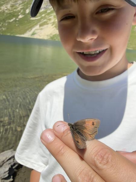 A young boy with a bright smile gently holds a butterfly on his finger, set against the serene backdrop of a mountain lake and green hills in Courchevel